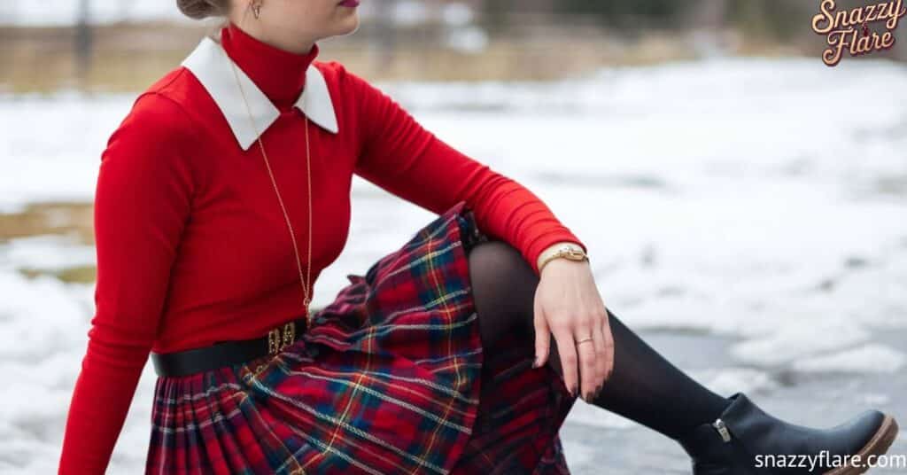 erson wearing a red top with a white collar, a plaid skirt, black tights, and black boots, sitting on a snowy ground