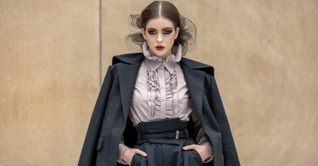 A model wearing a ruffled shirt, high-waisted trousers, and a structured jacket, with dramatic makeup and a ruffled hairstyle.